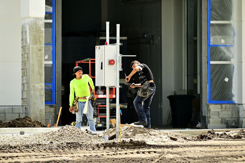 Two construction workers install an electrical panel outside a building site, wearing safety gear.