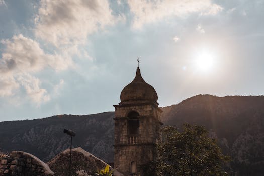 Scenic view of a historic bell tower against a mountainous backdrop with sunlight filtering through clouds.