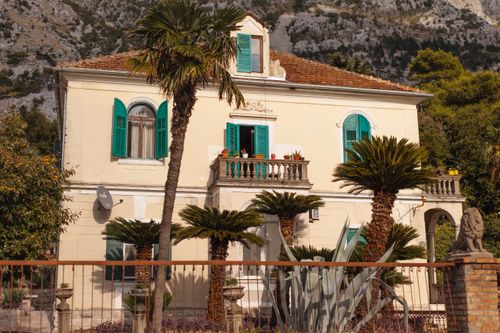 Free Beautiful Mediterranean villa with vibrant shutters, surrounded by palm trees in a mountainous setting. Stock Photo