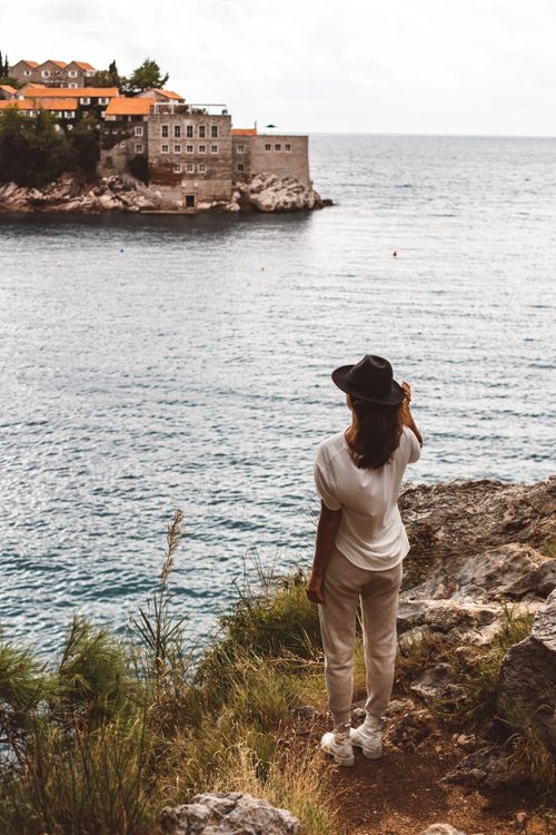 Free Woman standing on a rocky cliff gazing over the Adriatic Sea towards coastal buildings. Stock Photo