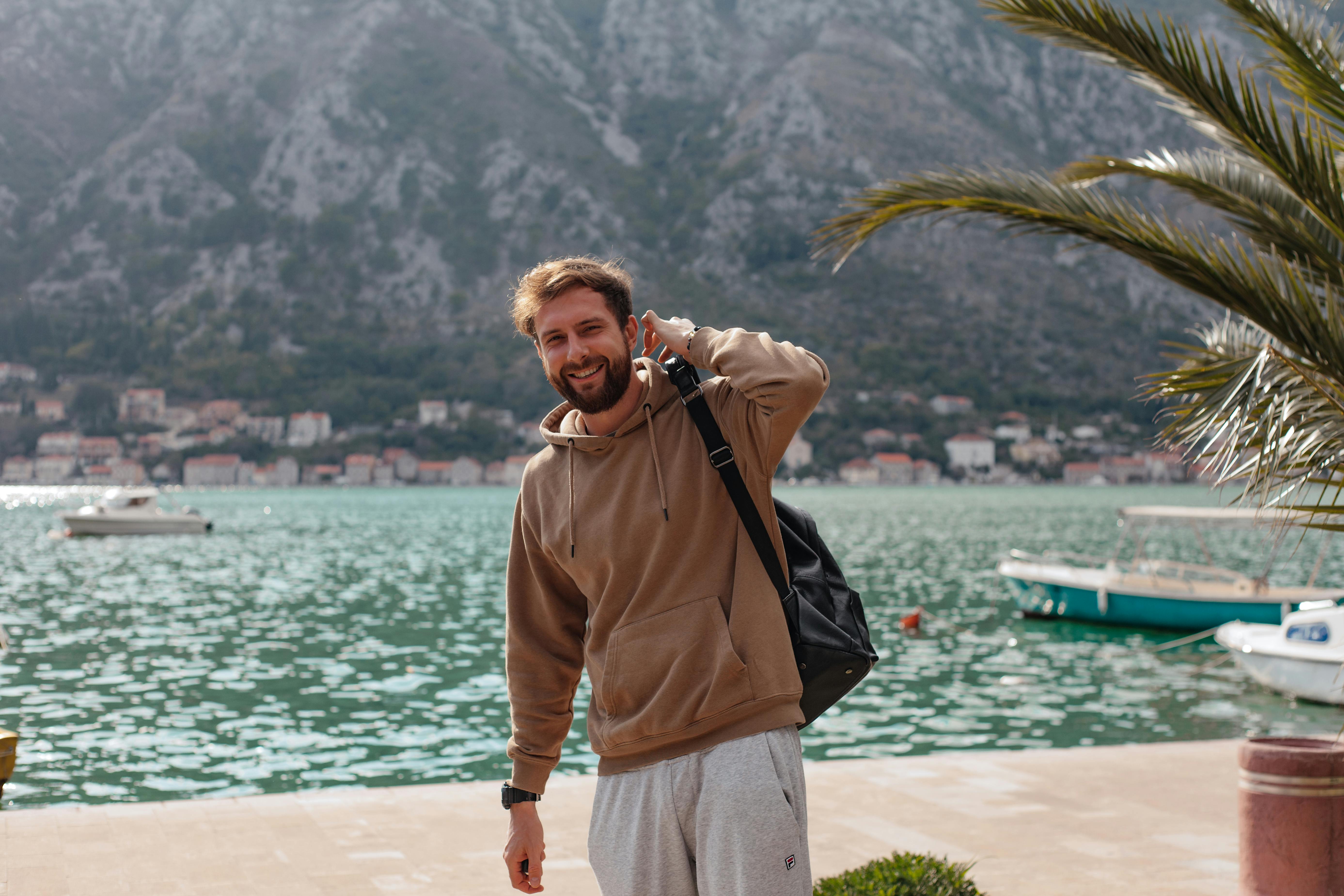 在风景如画的海岸背景下，一名穿着连帽衫的男子微笑着，周围有船只和山脉