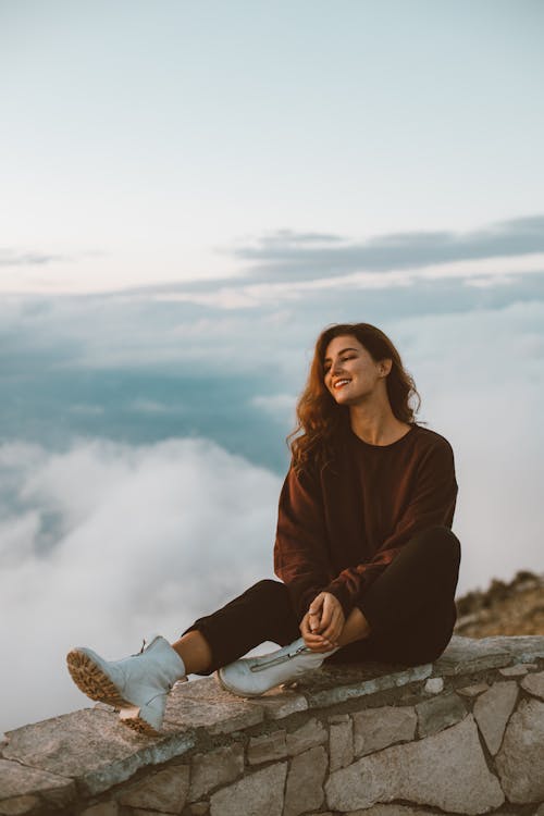 Woman in Brown Long Sleeve Shirt and Black Pants Sitting on Rock Fence