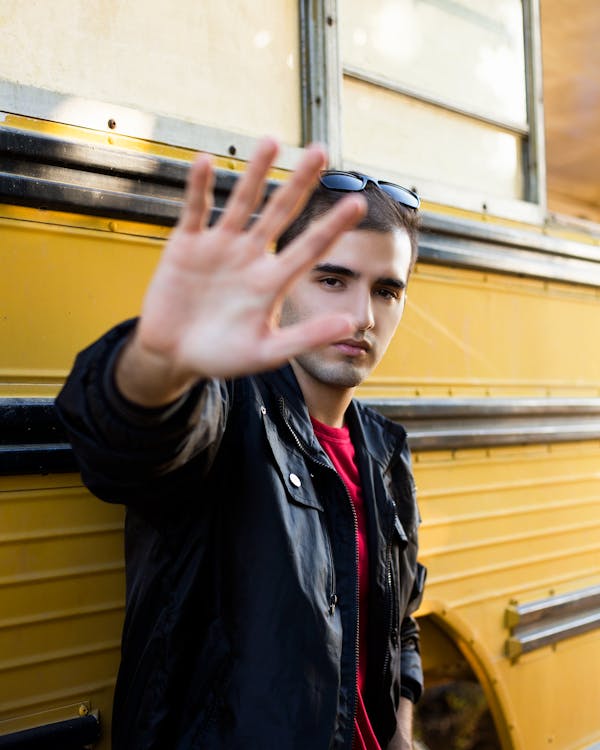Confident young male millennial in trendy outfit covering face with hand and looking at camera while standing near yellow bus