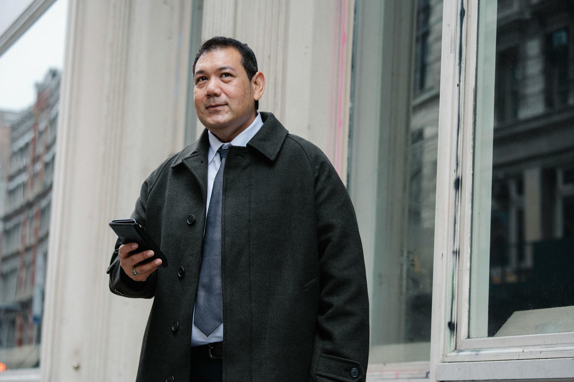 Low angle of confident adult ethnic male manager in classy suit and coat standing on street near building and browsing mobile phone