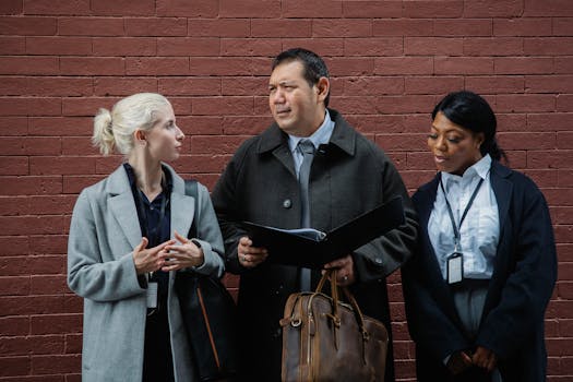 Concentrated multiracial coworkers in classic wear standing together near brick wall and checking documents in folder