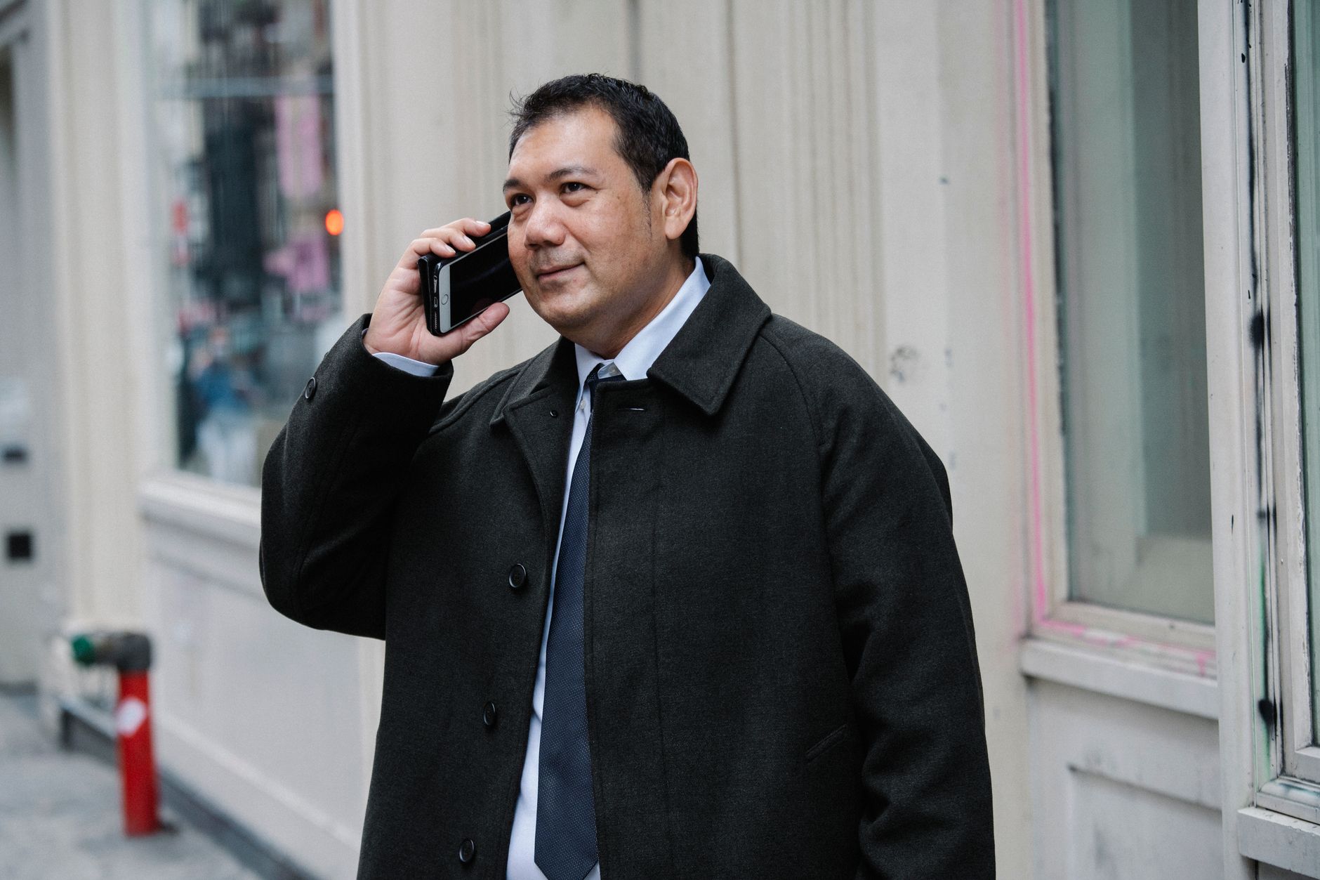 Positive adult ethnic male entrepreneur in classy white shirt and coat having phone conversation while standing on street
