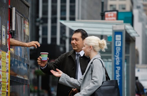 Free Side view of young female manager with ethnic male colleague in formal clothes buying takeaway coffee from booth on city street during break Stock Photo