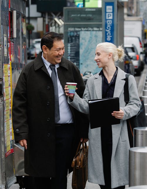 Free Cheerful diverse colleagues in classy outfits smiling and walking on city street while drinking takeaway coffee during break Stock Photo