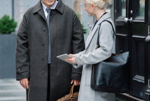 Crop woman in elegant coat with bag showing information on tablet to unrecognizable male colleague while standing together on street