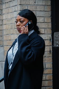 Elegant businesswoman in coat using smartphone on city street against a brick wall.