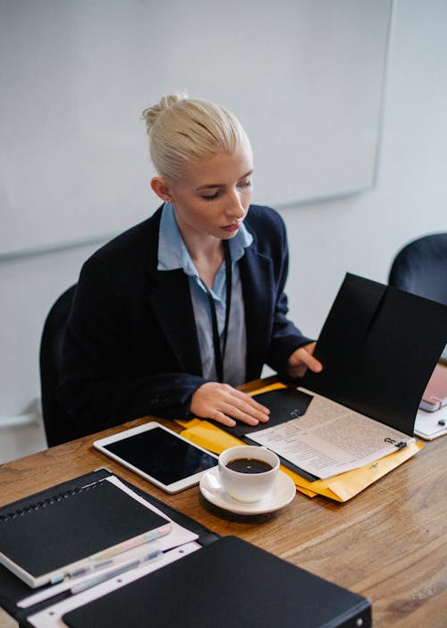 From above of focused young businesswoman in formal clothes reading documents placed in folder while working in office sitting at table with cup of coffee and tablet