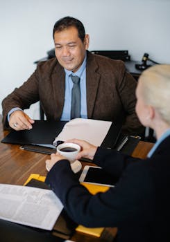 Two professionals in a meeting discussing documents over coffee at an office table.