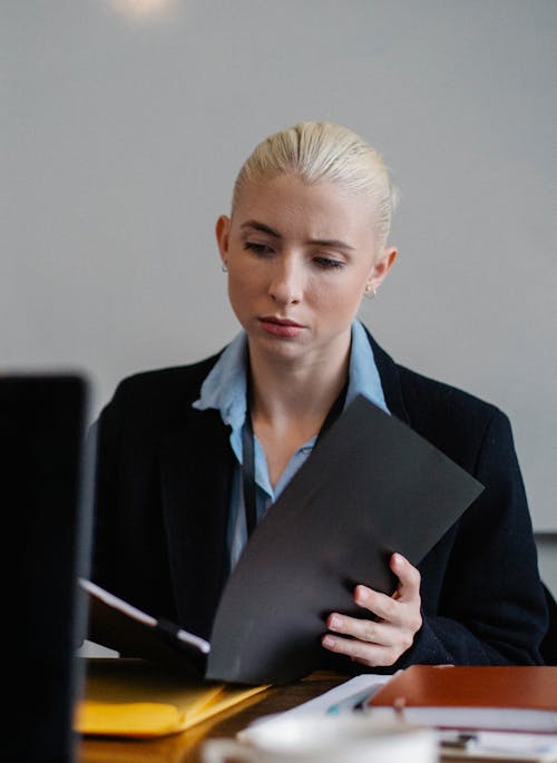 Free Serious young female employee with blond hair in formal suit sitting at table and reading documents with concentration in office Stock Photo