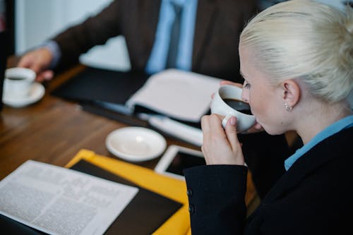 Side view of female in formal clothes drinking hot coffee while sitting at table with documents during business meeting