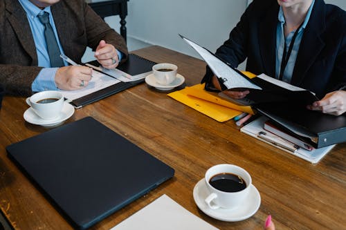 Crop unrecognizable coworkers in formal clothes sitting at table with documents and cups of coffee while working together
