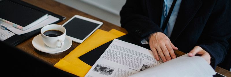 High angle of crop faceless businesswoman in formal clothes sitting at table with tablet and hot coffee and looking through documents