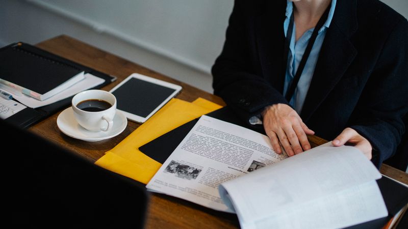 High angle of crop faceless businesswoman in formal clothes sitting at table with tablet and hot coffee and looking through documents