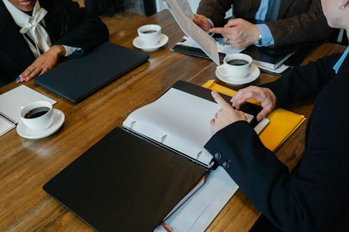 High angle of crop faceless coworkers in formal clothes sitting at wooden table with documents and fresh coffee while having meeting