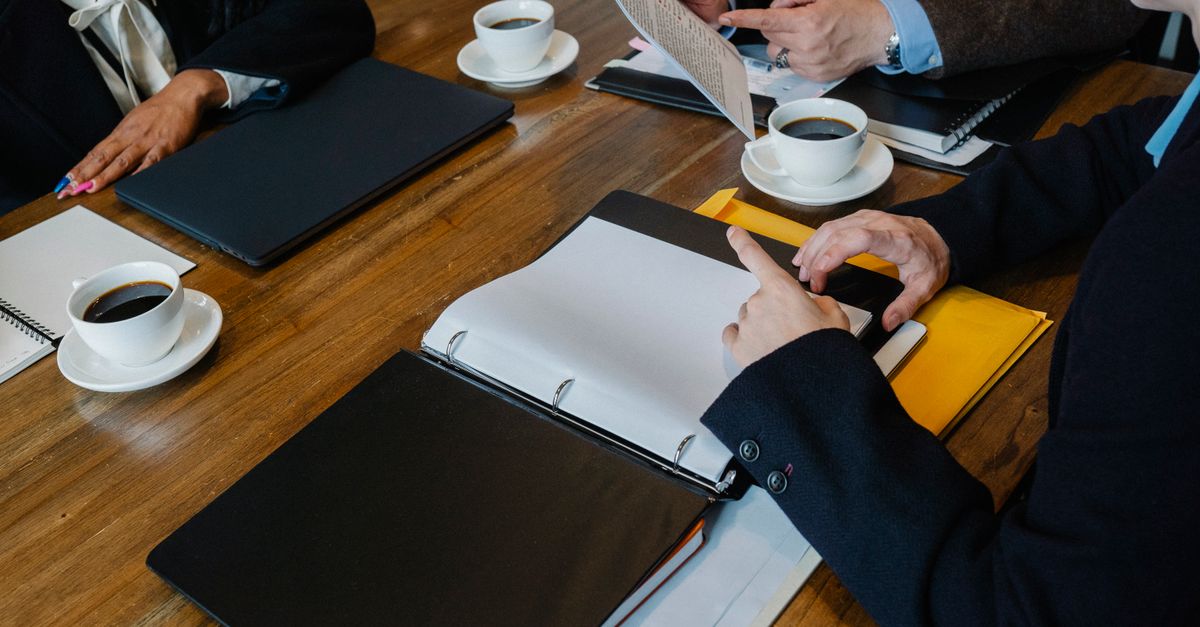 High angle of crop faceless coworkers in formal clothes sitting at wooden table with documents and fresh coffee while having meeting