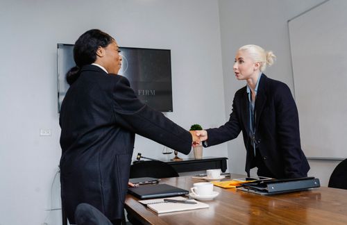 Free Smiling multiracial women in formal wear standing near table with documents and shaking hands after successful deal Stock Photo