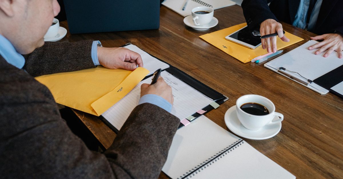 High angle of crop faceless coworkers in formal clothes sitting at wooden table with cups of coffee and documents while working together