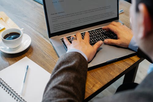 Crop faceless male entrepreneur in formal wear sitting at table with cup of coffee and notebook while working on project in laptop