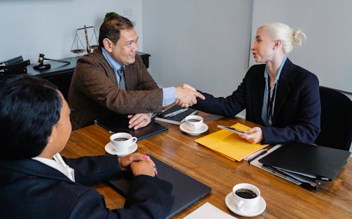 Businesspeople in formal wear sitting at table with documents and coffee while shaking hands after successful business deal