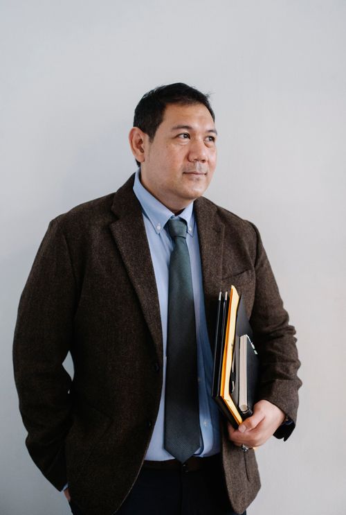 Free Positive adult ethnic male in formal suit standing with hand in pocket and with paper folders against white wall and looking away Stock Photo
