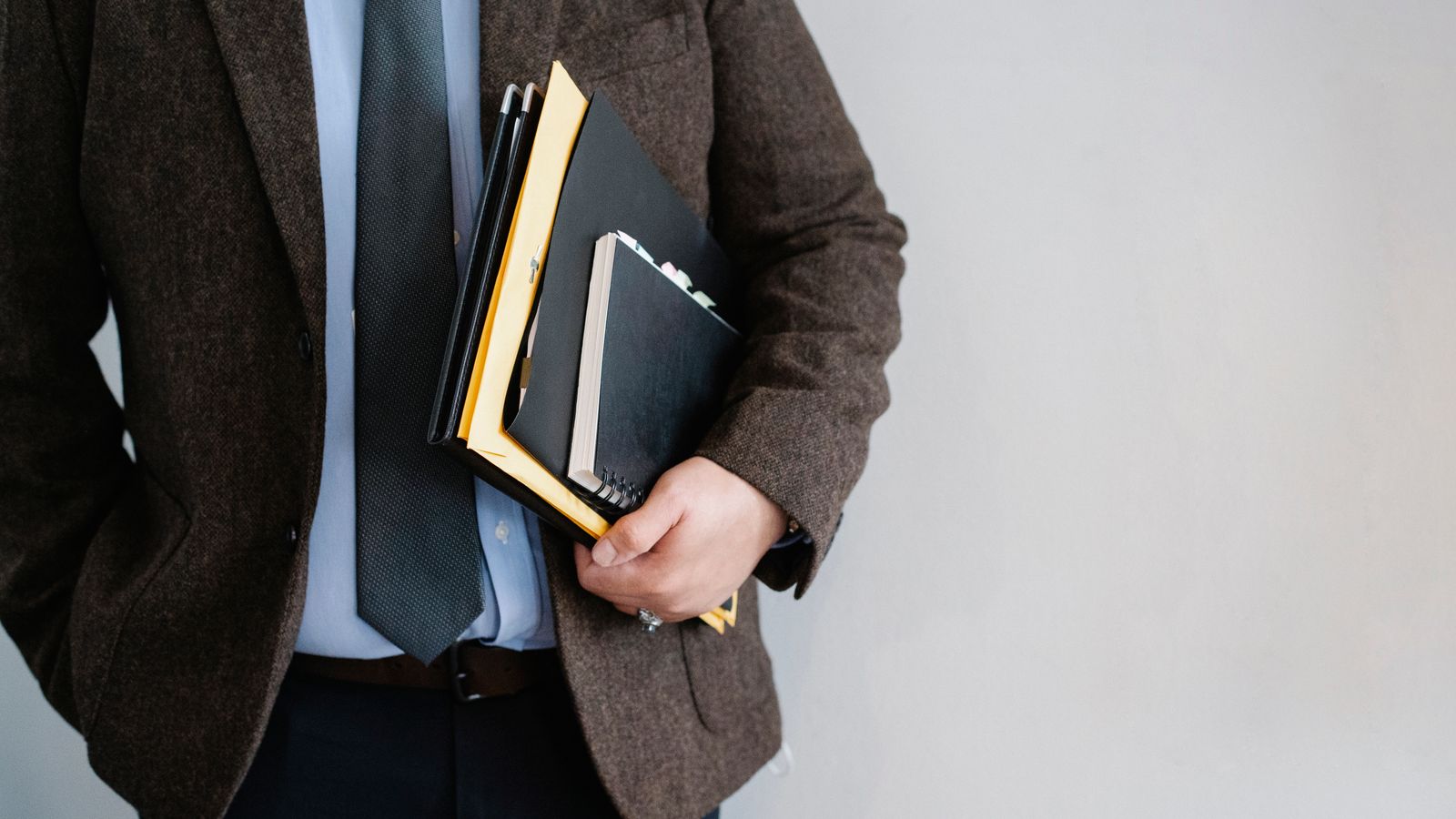 Crop anonymous male employee in formal suit standing with hand in pocket and paper folders in hand against white wall