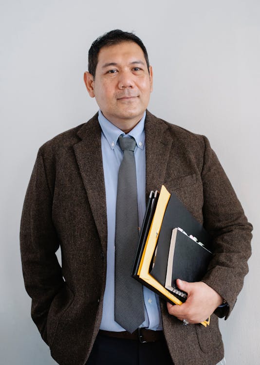 Free Positive Asian male office worker in formal suit with paper folders in hand standing with hand in pocket against white wall Stock Photo