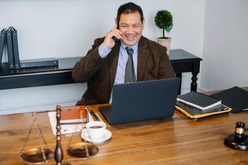 Free Joyful Asian male lawyer wearing formal suit having conversation on mobile phone and using laptop while working in modern law firm Stock Photo