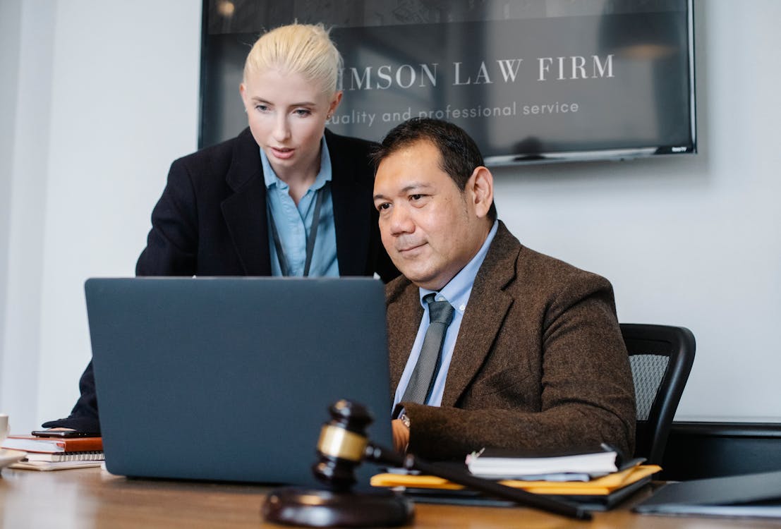 Smart mature ethnic judge in suit working on laptop and discussing details of new case with young female assistant in law firm