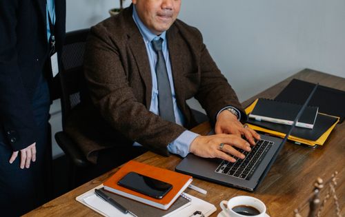 Free Crop anonymous ethnic male manager in suit and tie sitting at desk and typing on laptop while explaining job to unrecognizable colleague Stock Photo