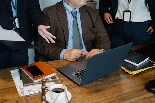 Free Crop faceless diverse colleagues in formal suits gathering around table with laptop and stack of  documents and negotiating about business strategy Stock Photo