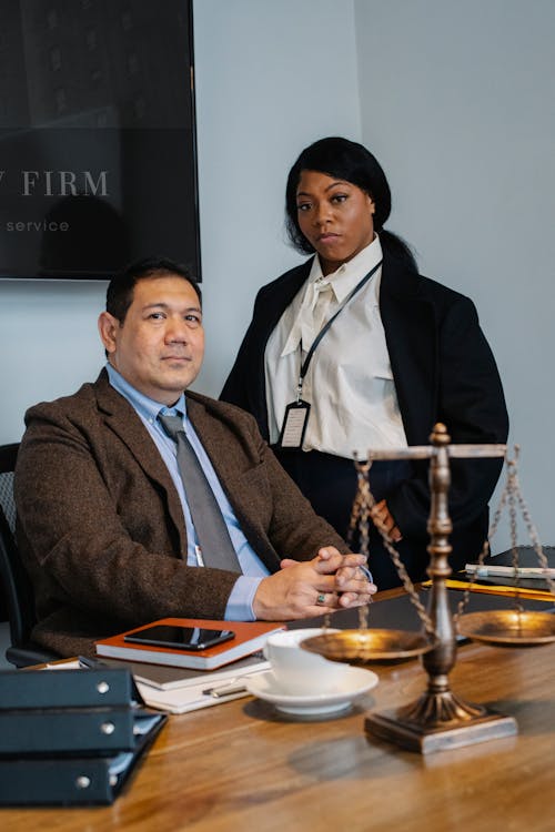 Focused young black woman in formal clothes standing near male ethnic colleague at wooden table with documents and justice scale during work in law firm