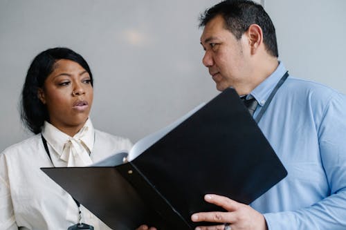 Serious middle aged male manager in shirt showing folder with documents to attentive female African American colleague during work in modern office