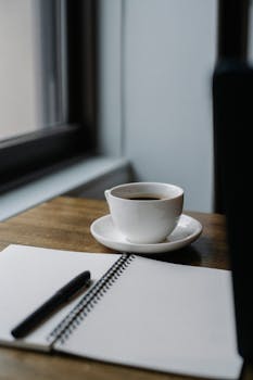 A cup of coffee and an open notebook by a window in a cozy café setting.