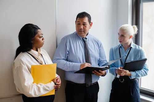 Free Group of diverse coworkers standing with documents near wall and discussing details of project in daytime Stock Photo