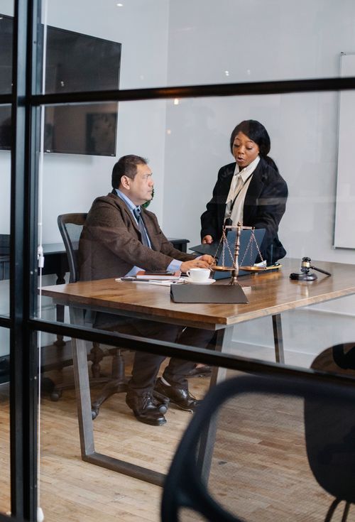 Free View through glass of serious black woman in formal suit explaining opinion to interested mature colleague in light office with judge gavel and scales on table Stock Photo