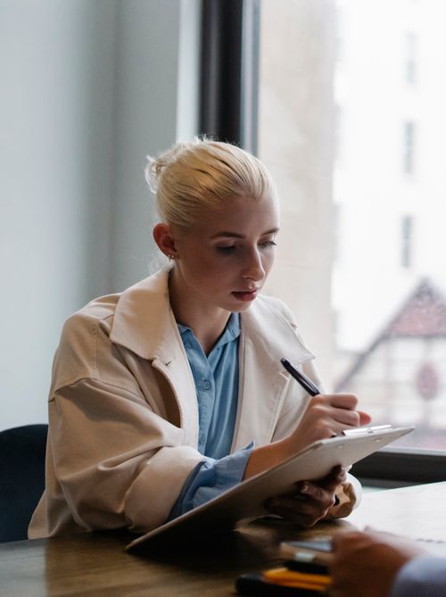 Free Young serious female with blond hair wearing elegant coat sitting at desk and writing ideas of business partner on clipboard in light office Stock Photo