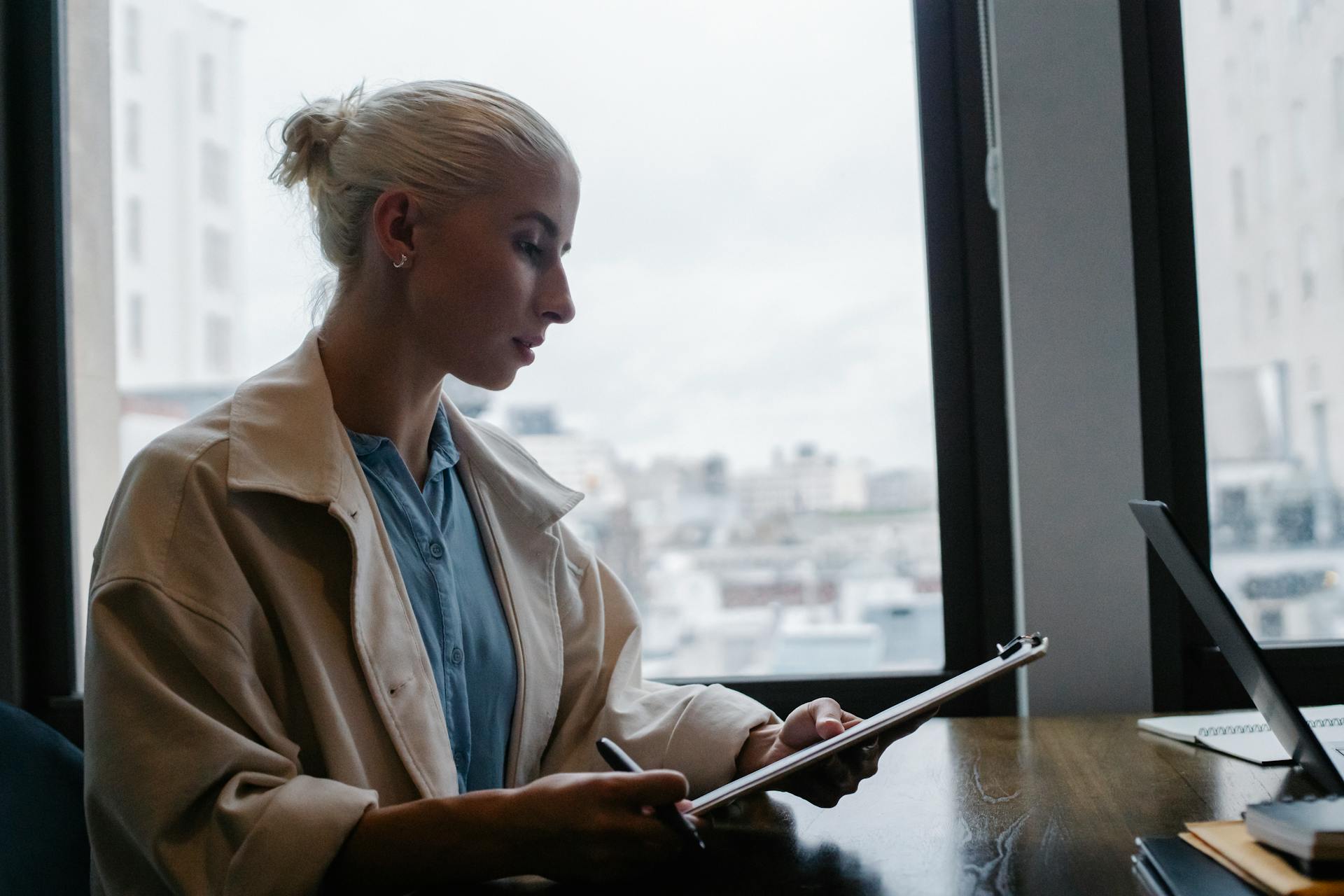 Side view of young concentrated female wearing white coat sitting at wooden desk in office and checking reports on clipboard