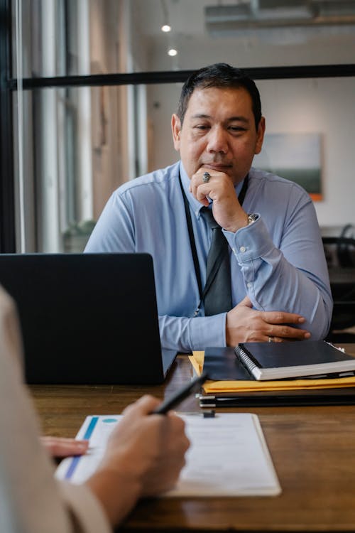Serious mature ethnic entrepreneur sitting at wooden table with documents and laptop and thinking while crop anonymous colleague taking notes on clipboard