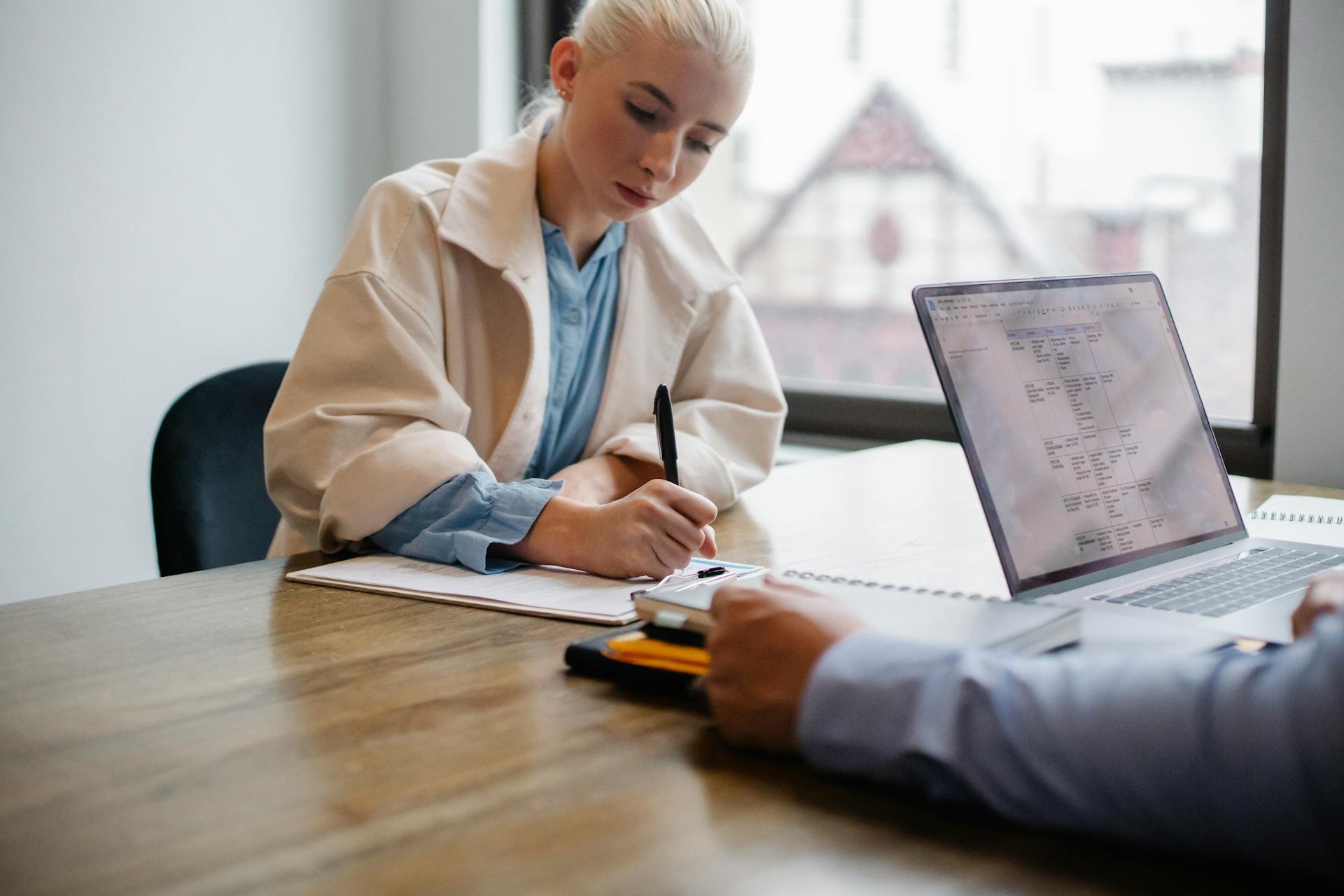 Serious young female manager in formal outfit sitting and signing documents while business partner working on laptop