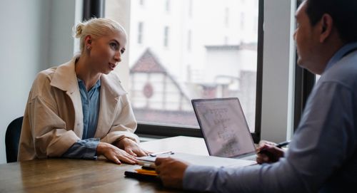 Free Crop man sitting at wooden desk with laptop and documents and listening attentively to positive young woman in trendy outfit in light workspace Stock Photo