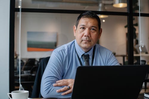 Confident ethnic male entrepreneur using laptop during business work in modern workspace and looking at camera