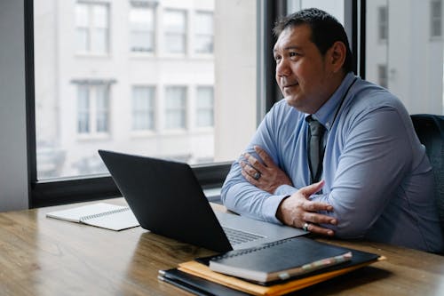 Free Adult successful ethnic male boss wearing shirt and tie sitting with hands crossed at workplace with documents and netbook Stock Photo