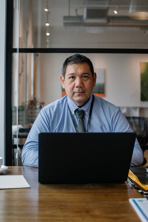 Confident ethnic man wearing formal clothes and tie working at netbook while sitting at table in workspace