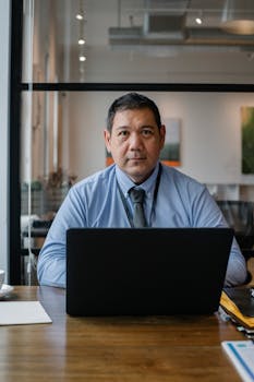 Confident ethnic man wearing formal clothes and tie working at netbook while sitting at table in workspace