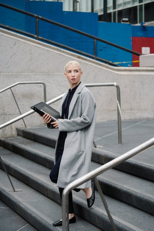 Full body of serious woman wearing long coat carrying folder with documents and notebook in office building