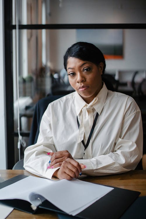 Serious African American businesswoman wearing formal white blouse and sitting at office desk with documents while looking at camera